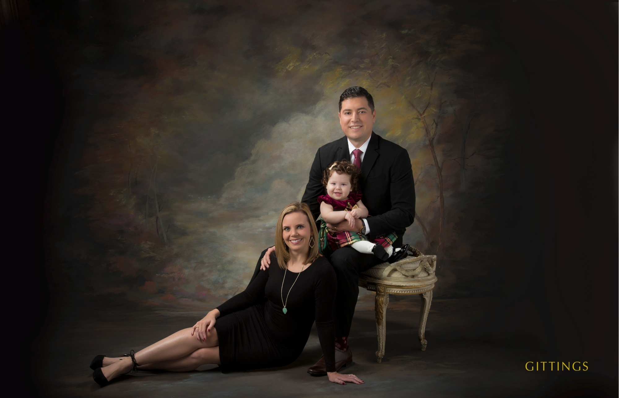 Studio Family Portrait Of Parents And Their Young Daughter, Dressed In Classic Black And Deep Tones Against A Painted Backdrop.